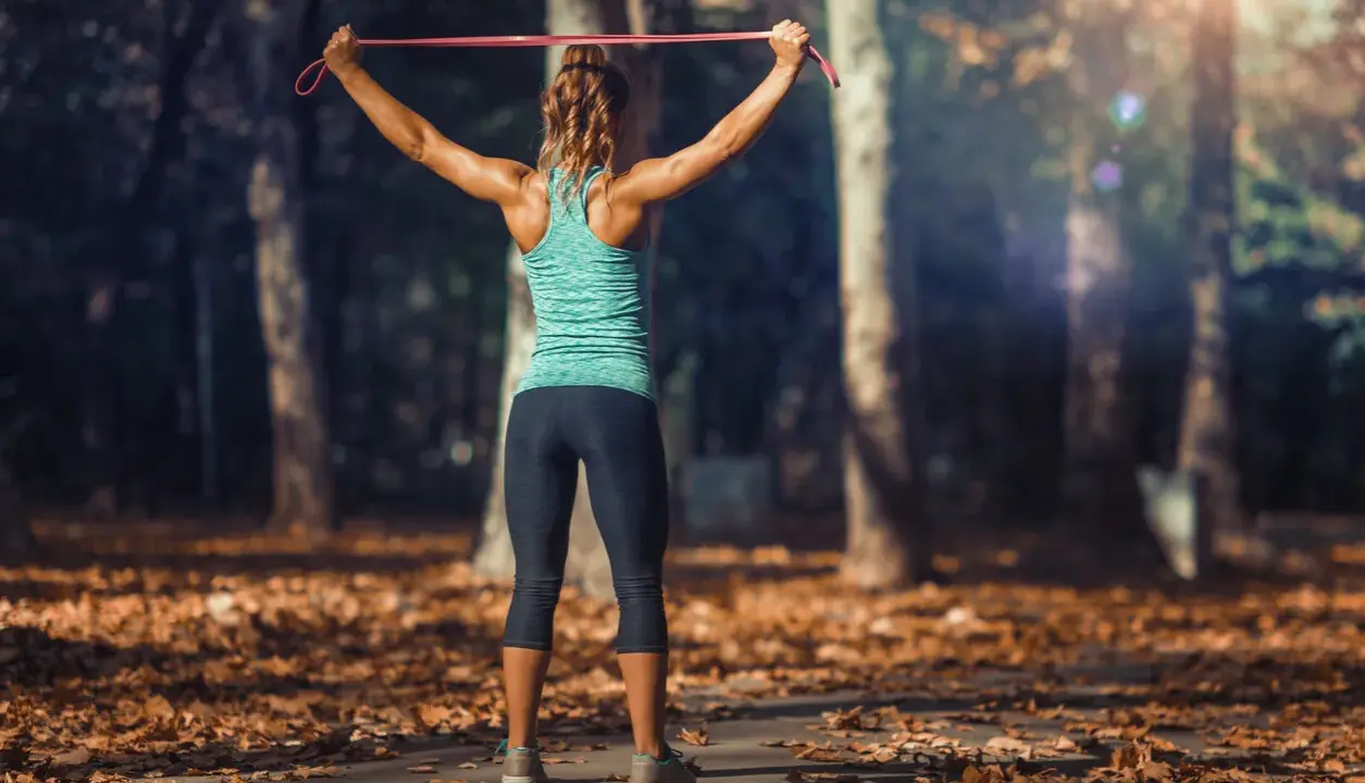 Mujer entrenando tren superior con tabla de ejercicios y gomas elásticas en un parque otoñal.