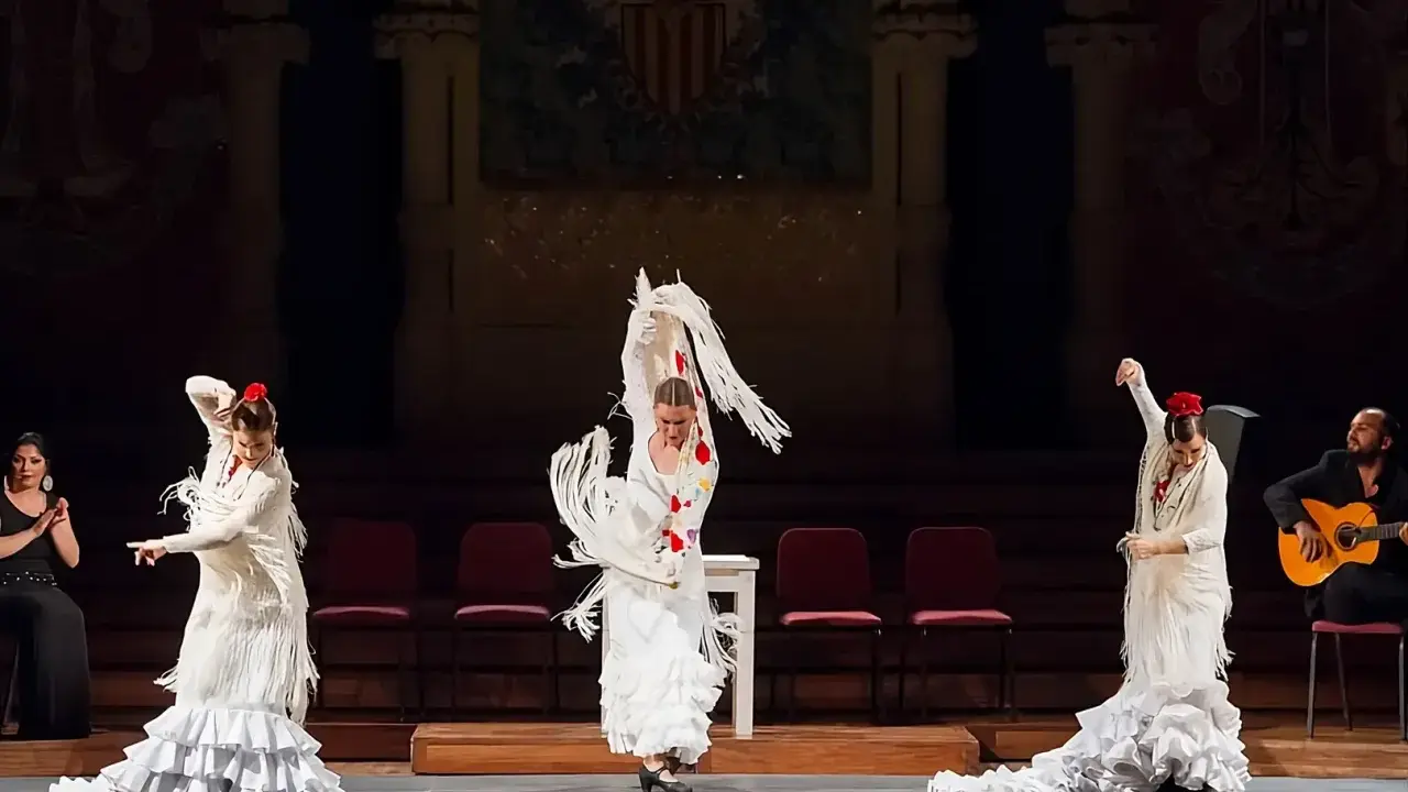 Tres bailarinas de flamenco, con trajes blancos y volantes, ejecutan un **taconeo flamenco** en un escenario. Un guitarrista acompaña la actuación.