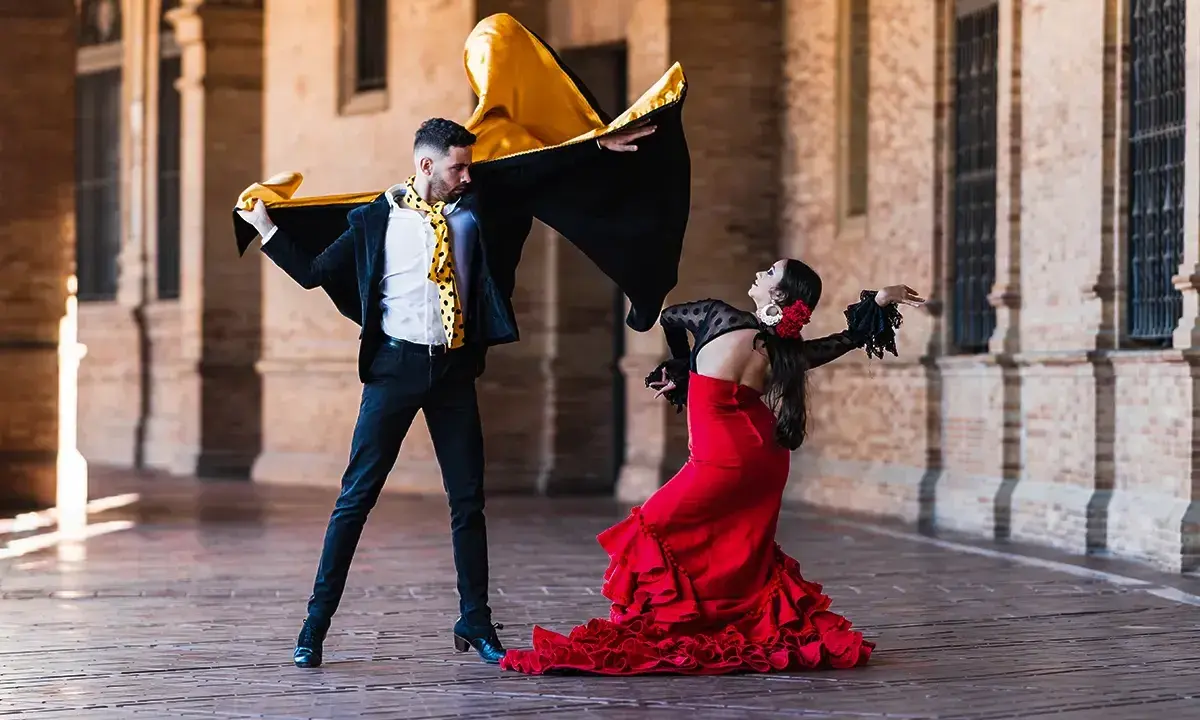 Pareja bailando flamenco con pasión. Él, con capa negra y dorada, ella, con vestido rojo voluminoso. Un jaleo flamenco lleno de arte.