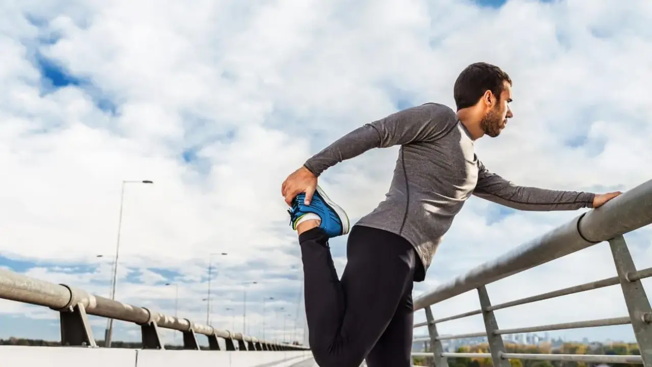 Hombre haciendo estiramientos antes de correr en un puente, con el cielo nublado de fondo.