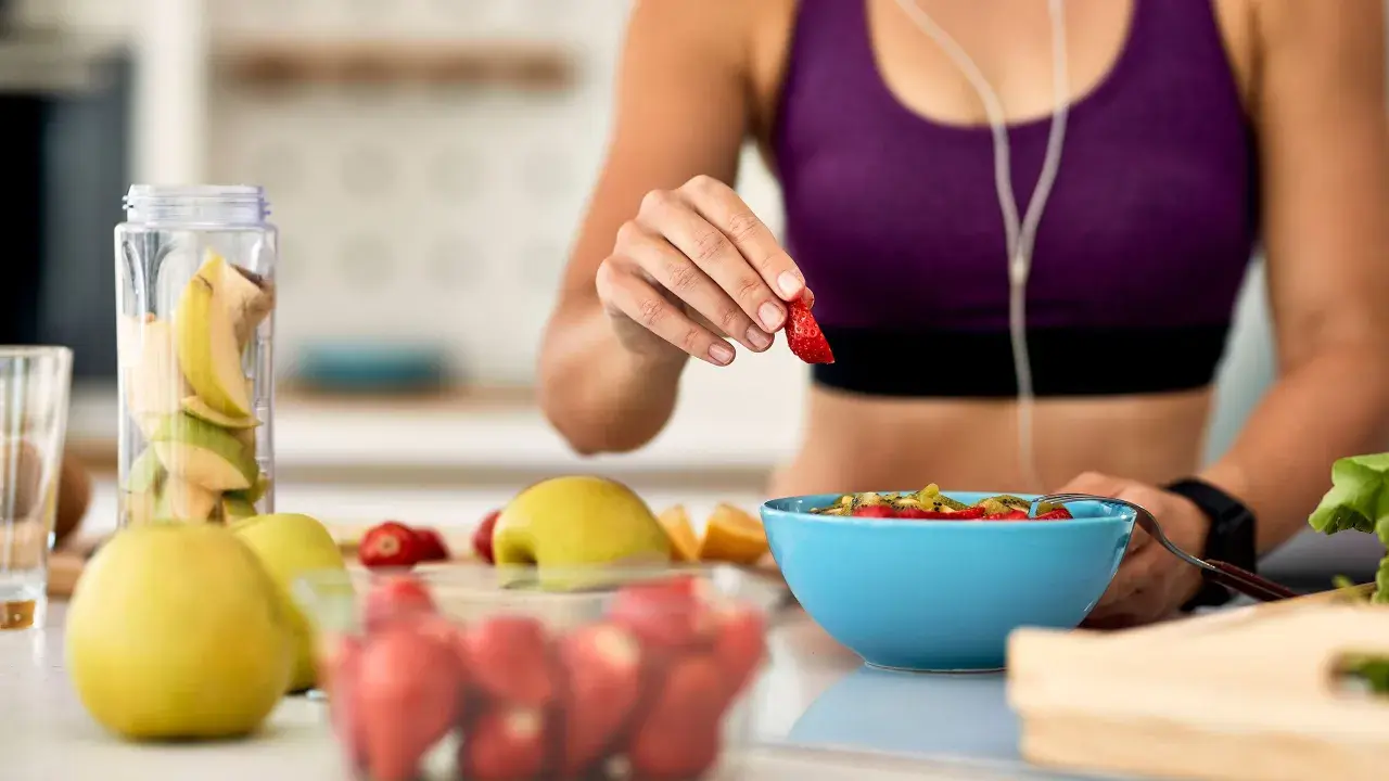 Mujer en ropa deportiva prepara su desayuno saludable con frutas frescas, pensando en qué desayunar antes de entrenar.