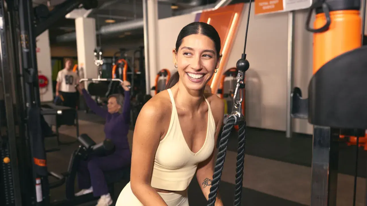 Mujer sonríe en gimnasio, mientras otros realizan ejercicios compuestos y aislados en máquinas.