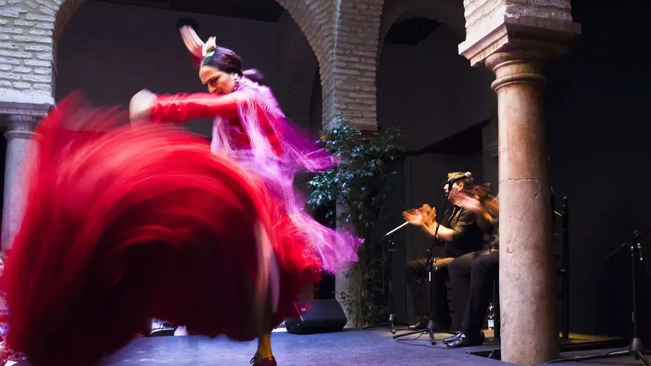 Una bailaora española gira con su vestido rojo, creando un torbellino de tela. A su lado, un guitarrista acompaña con su música.