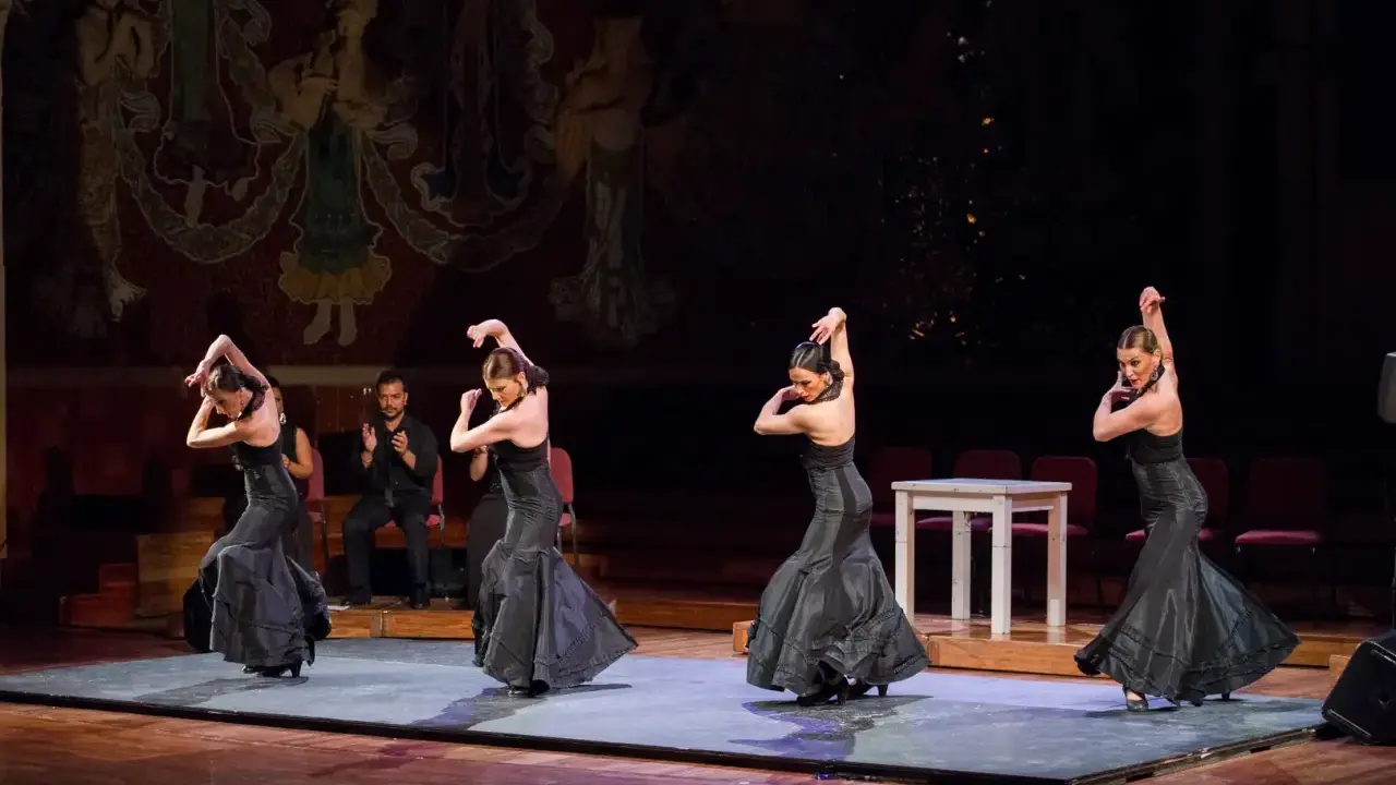 Cuatro bailarinas de flamenco, vestidas de negro, ejecutan pasos apasionados. Un percusionista las acompaña.