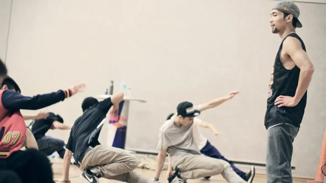 Jóvenes practicando una coreografía de break dance, con movimientos en el suelo y un instructor observando.