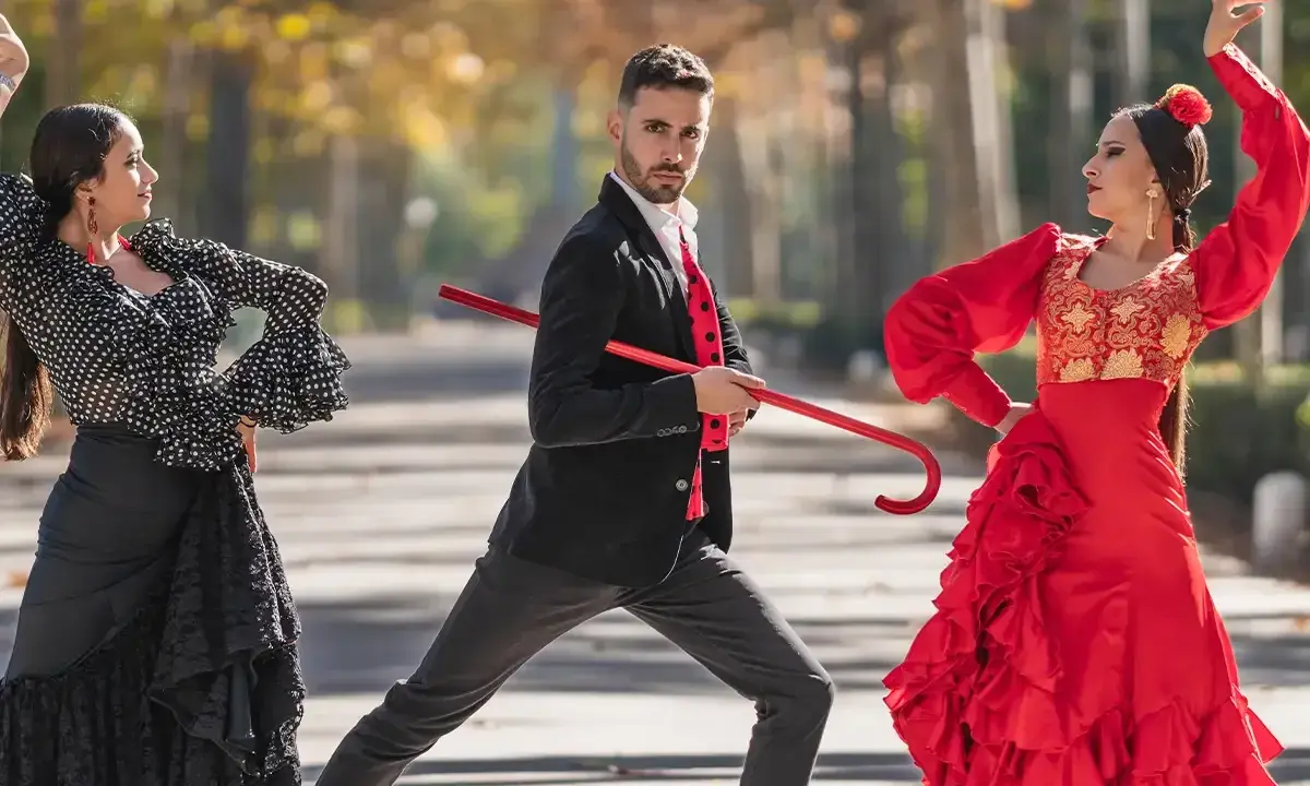 Bailarines de flamenco, uno con traje negro y rojo, sostiene un bastón rojo, mientras dos mujeres con trajes vibrantes ejecutan pasos. Explora los tipos de palos flamencos.