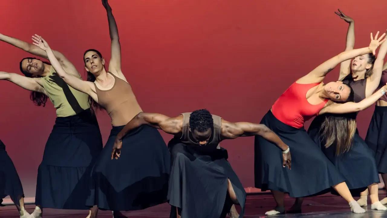 Un grupo de bailarines realiza movimientos fluidos en unas clases de danza, con faldas negras y un fondo rojo vibrante.