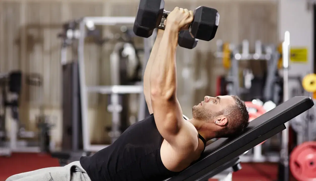 Hombre en gimnasio haciendo press de banca con mancuernas. Un ejercicio clave en una tabla de entrenamiento para ganar masa muscular y perder grasa.