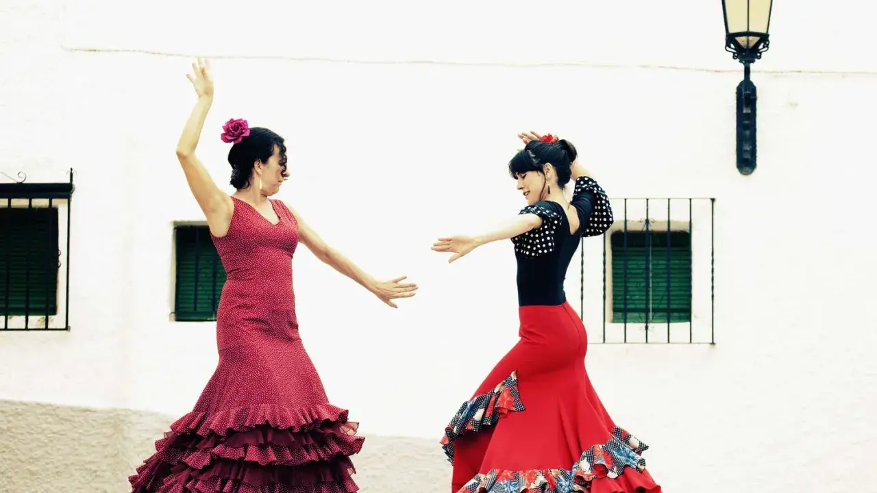 Dos mujeres con trajes vibrantes ejecutan apasionados bailes flamencos frente a una pared blanca.