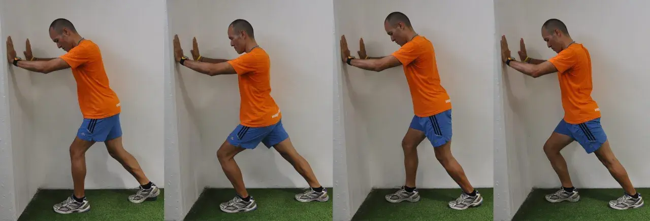 Hombre realizando estiramientos después de correr contra una pared, apoyando las manos y extendiendo una pierna hacia atrás para estirar el gemelo.