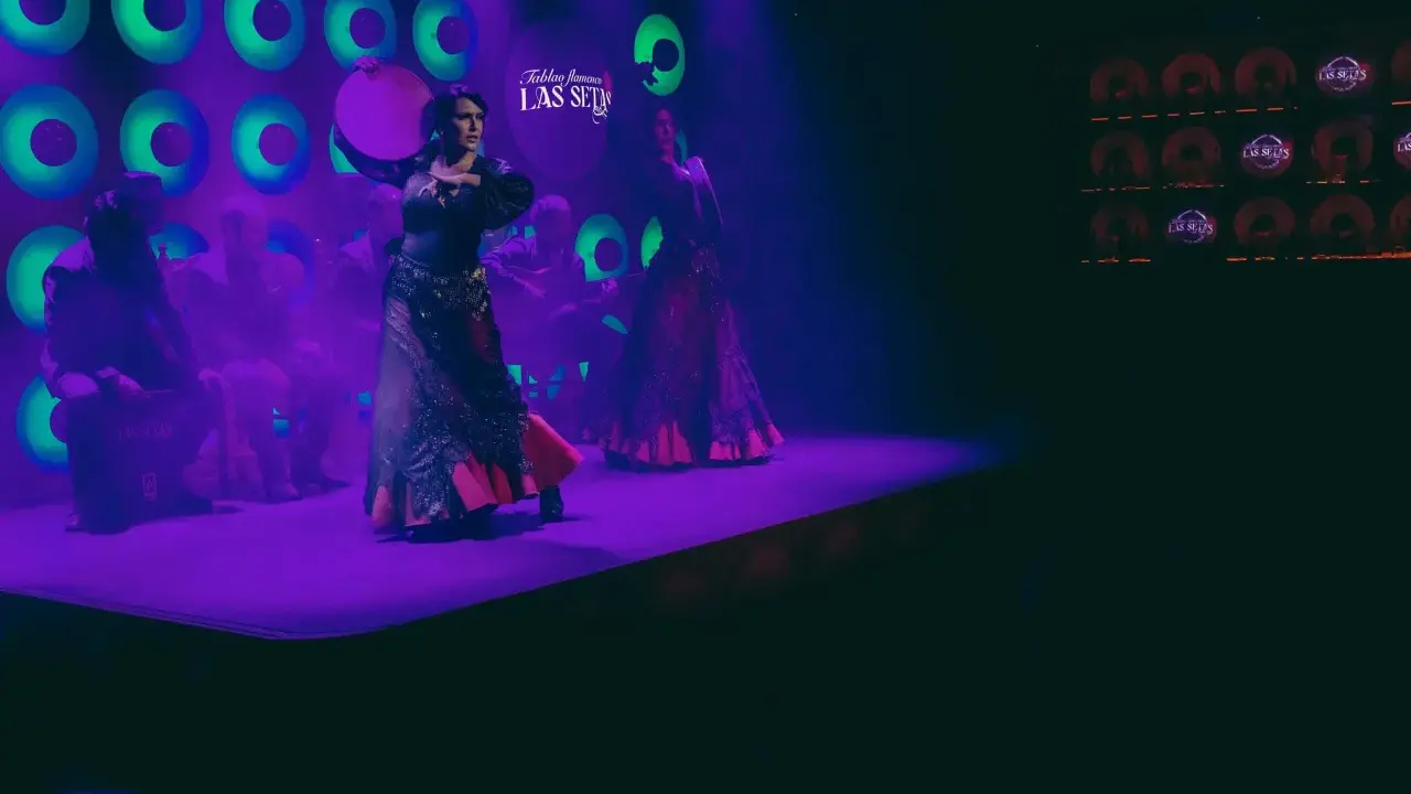 Dos bailarinas de danza sevillana, con trajes vibrantes, actúan bajo luces púrpuras y verdes.
