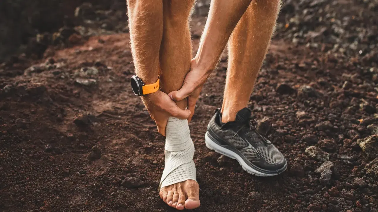 Corredor con tobillo vendado, sintiendo dolor espinilla al correr. Reloj deportivo naranja en muñeca.