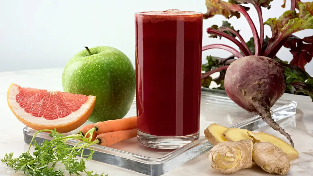 Vaso de zumo de remolacha vibrante rodeado de ingredientes frescos: remolacha, zanahorias, manzana verde, pomelo y jengibre.