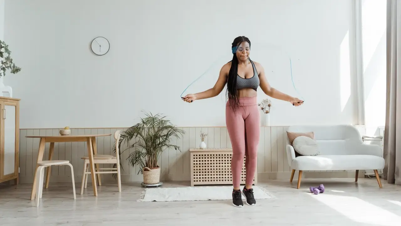 Mujer joven con auriculares realiza ejercicio hit saltando la cuerda en su sala de estar.