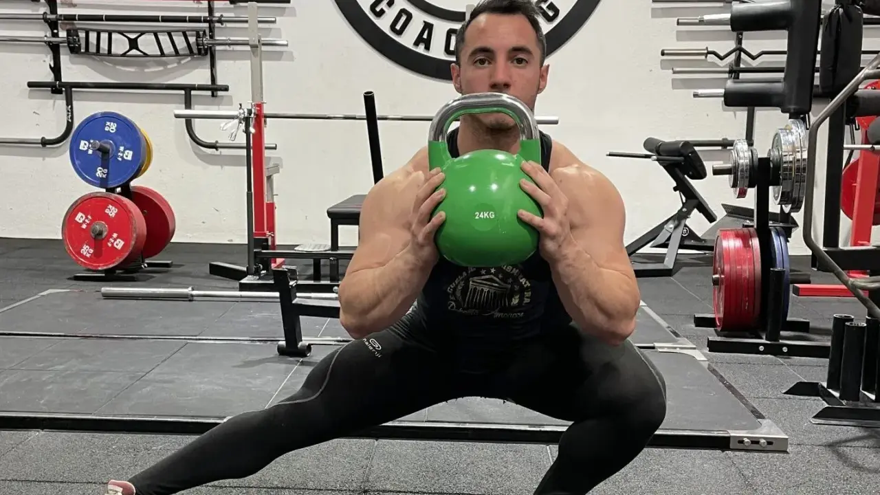 Hombre realiza desplantes laterales con pesa rusa verde en un gimnasio.