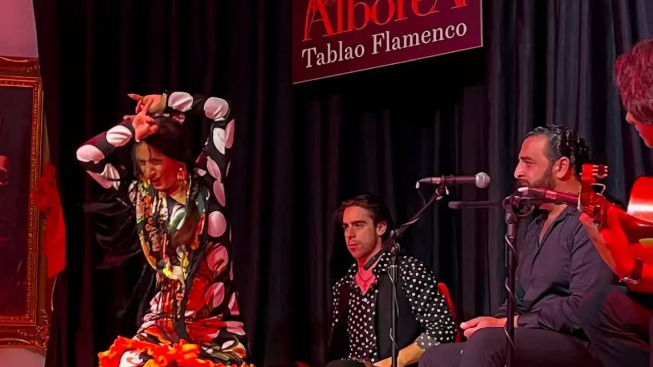 Una bailaora de flamenco, con un vestido de lunares y volantes, actúa en un tablao. A su lado, músicos la acompañan con guitarra y cante.