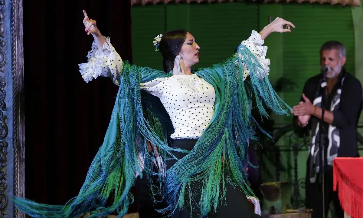Bailarina de flamenco con mantón verde vibrante, sus movimientos evocan frases flamenco. Un hombre aplaude al fondo.