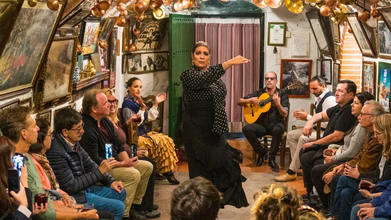 Una bailaora de flamenco, con un vestido negro de lunares, actúa en un local íntimo donde se baila flamenco. Un guitarrista la acompaña.