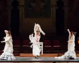 Tres bailarinas de flamenco, con trajes blancos y volantes, ejecutan un **taconeo flamenco** en un escenario. Un guitarrista acompaña la actuación.