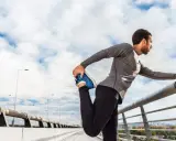 Hombre haciendo estiramientos antes de correr en un puente, con el cielo nublado de fondo.