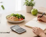 Calculadora y plato de comida saludable junto a una persona anotando en un cuaderno, para saber cuantas calorias necesito.