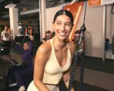 Mujer sonríe en gimnasio, mientras otros realizan ejercicios compuestos y aislados en máquinas.