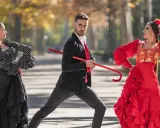Bailarines de flamenco, uno con traje negro y rojo, sostiene un bastón rojo, mientras dos mujeres con trajes vibrantes ejecutan pasos. Explora los tipos de palos flamencos.