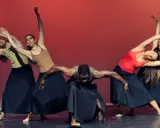 Un grupo de bailarines realiza movimientos fluidos en unas clases de danza, con faldas negras y un fondo rojo vibrante.