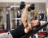 Hombre en gimnasio haciendo press de banca con mancuernas. Un ejercicio clave en una tabla de entrenamiento para ganar masa muscular y perder grasa.
