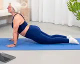 Mujer mayor con pelo blanco realiza postura de cobra en una esterilla azul, parte de su entrenamiento para mujeres.