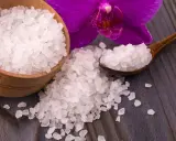 Sal de Epsom en un cuenco de madera y cuchara, junto a una orquídea morada. Descubre para qué sirve la sal de Epsom para tu bienestar.