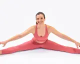 Mujer sonriente en ropa deportiva rosa, practicando una postura de yoga que demuestra diferentes tipos de flexibilidad, con las piernas abiertas y tocando los dedos de los pies.