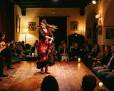 Una bailaora de flamenco en vivo, con un vestido rojo y negro, gira en un escenario de madera. Dos guitarristas la acompañan mientras el público observa.