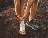 Corredor con tobillo vendado, sintiendo dolor espinilla al correr. Reloj deportivo naranja en muñeca.