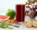 Vaso de zumo de remolacha vibrante rodeado de ingredientes frescos: remolacha, zanahorias, manzana verde, pomelo y jengibre.