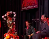 Una bailaora de flamenco, con un vestido de lunares y volantes, actúa en un tablao. A su lado, músicos la acompañan con guitarra y cante.
