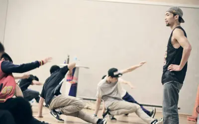 Jóvenes practicando una coreografía de break dance, con movimientos en el suelo y un instructor observando.