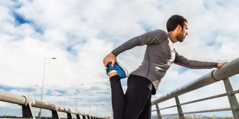 Hombre haciendo estiramientos antes de correr en un puente, con el cielo nublado de fondo.