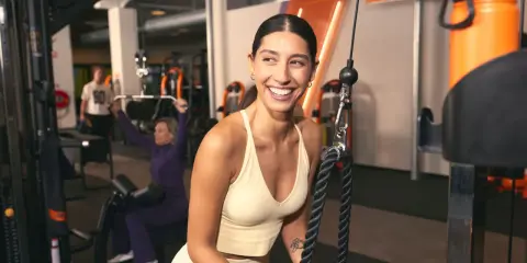 Mujer sonríe en gimnasio, mientras otros realizan ejercicios compuestos y aislados en máquinas.