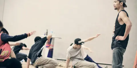 Jóvenes practicando una coreografía de break dance, con movimientos en el suelo y un instructor observando.