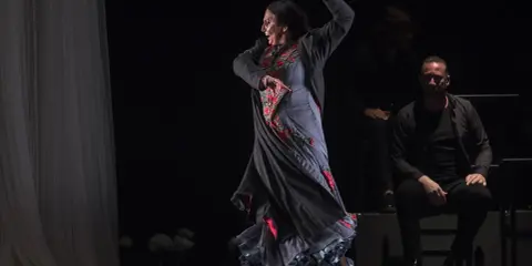Una bailaora de flamenco en pleno movimiento, con su falda ondeando y un hombre sentado observando.