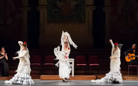 Tres bailarinas de flamenco, con trajes blancos y volantes, ejecutan un **taconeo flamenco** en un escenario. Un guitarrista acompaña la actuación.