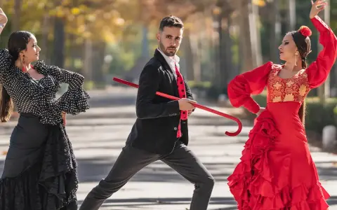 Bailarines de flamenco, uno con traje negro y rojo, sostiene un bastón rojo, mientras dos mujeres con trajes vibrantes ejecutan pasos. Explora los tipos de palos flamencos.