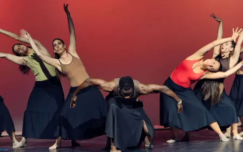 Un grupo de bailarines realiza movimientos fluidos en unas clases de danza, con faldas negras y un fondo rojo vibrante.