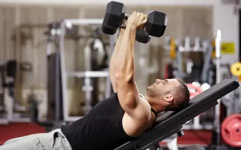 Hombre en gimnasio haciendo press de banca con mancuernas. Un ejercicio clave en una tabla de entrenamiento para ganar masa muscular y perder grasa.