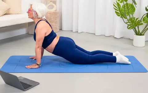 Mujer mayor con pelo blanco realiza postura de cobra en una esterilla azul, parte de su entrenamiento para mujeres.