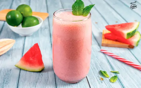 Refrescante zumo de sandía en vaso alto, decorado con menta. Alrededor, trozos de sandía, limones y una pajita a rayas.
