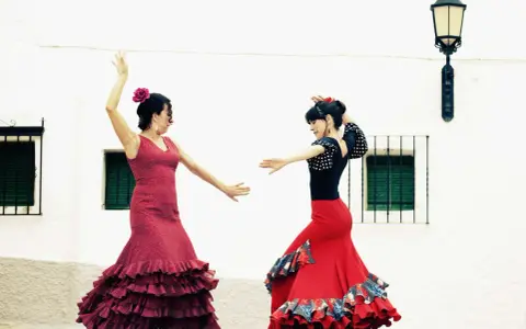 Dos mujeres con trajes vibrantes ejecutan apasionados bailes flamencos frente a una pared blanca.