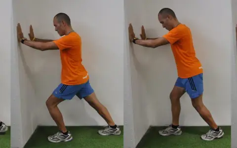 Hombre realizando estiramientos después de correr contra una pared, apoyando las manos y extendiendo una pierna hacia atrás para estirar el gemelo.
