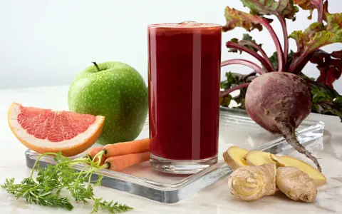 Vaso de zumo de remolacha vibrante rodeado de ingredientes frescos: remolacha, zanahorias, manzana verde, pomelo y jengibre.